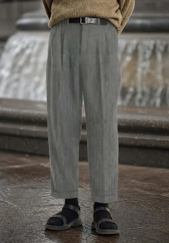 Front view of light gray pleated wool tweed dress pants with a cuffed hem, worn with a beige knit sweater, black belt, black socks, and dark sandals.
