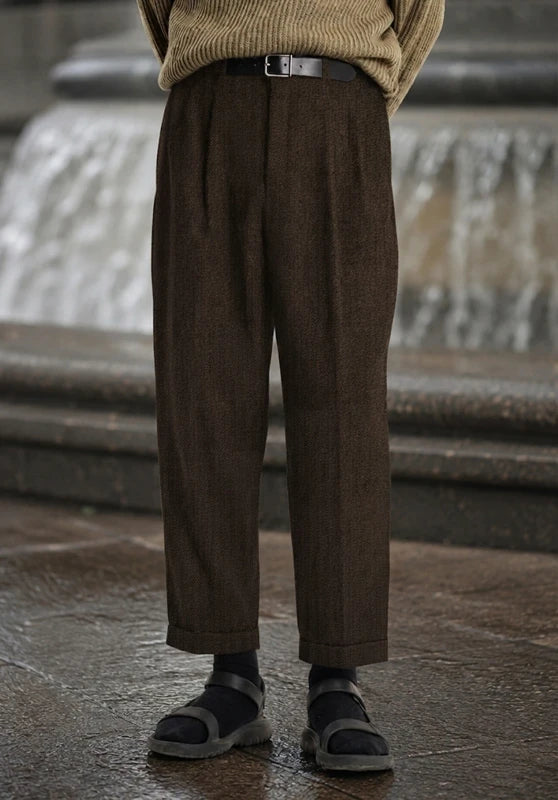 Front close-up view of dark brown pleated wool tweed dress pants featuring a belted waist, straight leg, and cuffed hem, styled with black socks and sandals against an outdoor fountain background.