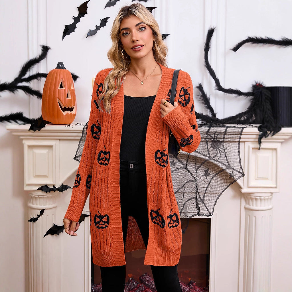 Full-length front view of a model standing in front of a fireplace wearing the orange open-front pumpkin knit longline cardigan over a black top and pants.