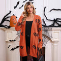 Three-quarter front view of a model in a Halloween-decorated room wearing an orange open-front longline ribbed knit cardigan with black jack-o'-lantern pumpkin motifs and dual front patch pockets.