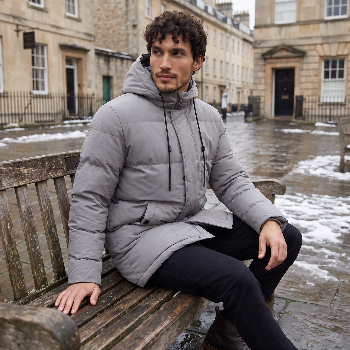 Gray longline quilted parka with a drawstring hood and concealed zip placket, worn by a man sitting on a wooden bench.
