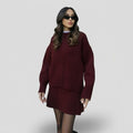 Three-quarter front view of a model wearing black sunglasses and a dark red relaxed knit set featuring an oversized crewneck sweater with dropped shoulders and a matching draping mini skirt, styled over sheer black tights.