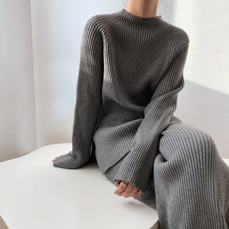 Model sitting relaxed on a white surface wearing the charcoal gry ribbed knit mock-neck sweater and wide-leg pants set.
