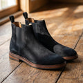 Black suede Chelsea boots with round toes, elastic side panels, and a slim welted sole on a rustic wooden floor.