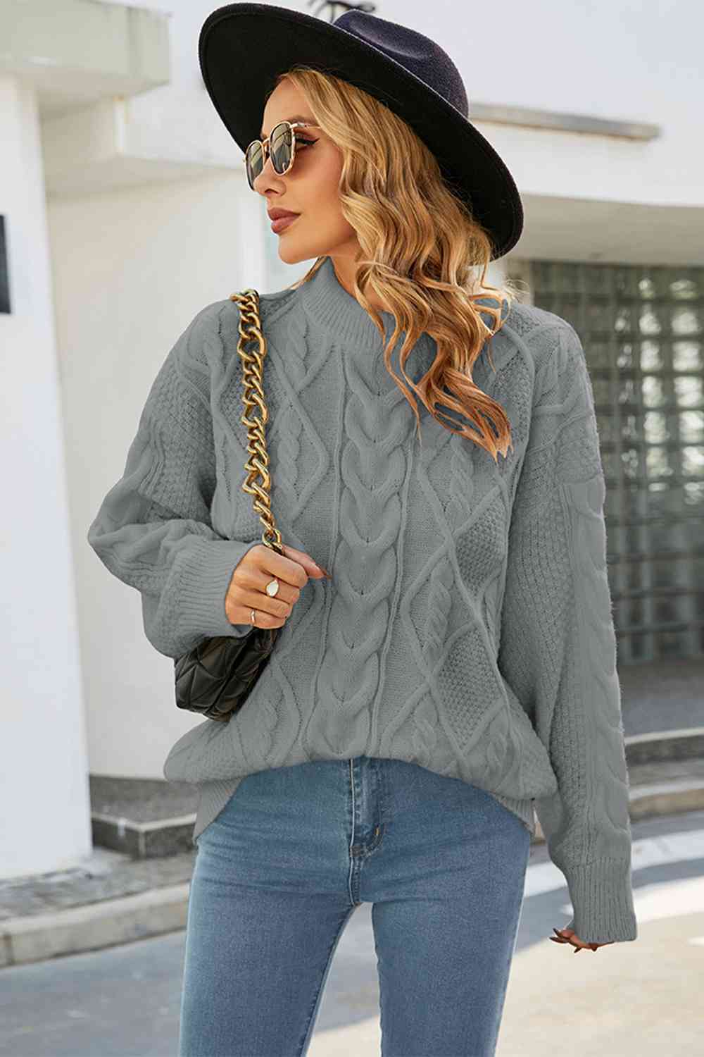 Grey cable-knit oversized crewneck sweater with drop shoulders and long sleeves, styled with a black hat and handbag.