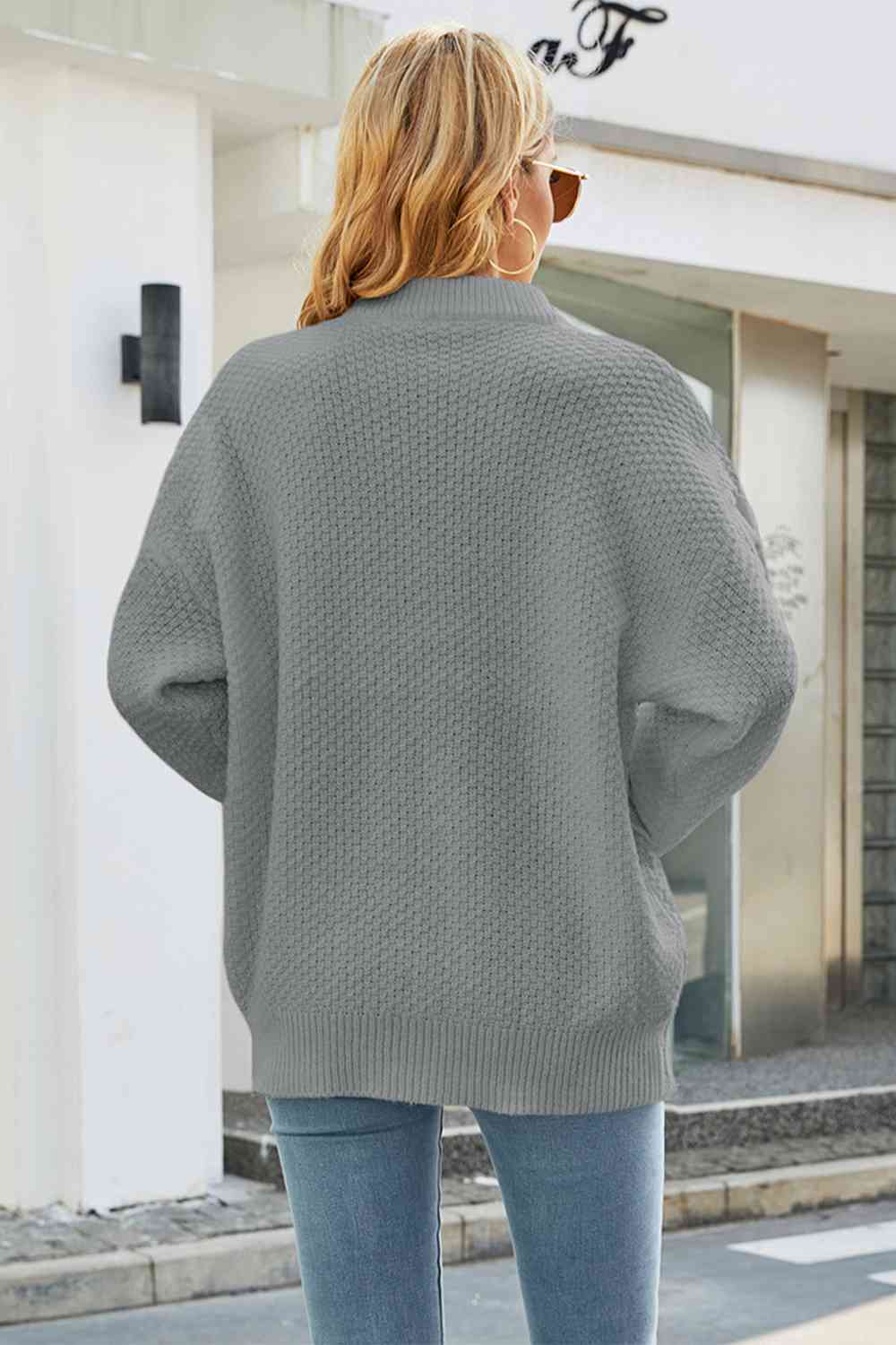 Grey oversized cable-knit sweater with a relaxed drop-shoulder fit and ribbed crewneck collar worn with blue jeans.