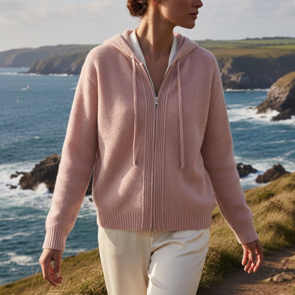 Soft pink ribbed cotton sweater coat with a drawstring hood and double-zip front, worn over white trousers by the coast.