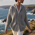 Grey ribbed cotton hooded sweater coat with a double-zip front, worn over white trousers by the coast.