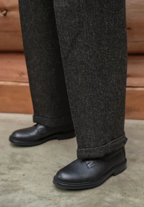 Black herringbone wool tweed trousers with a straight-leg fit and cuffed hems worn with black leather shoes.