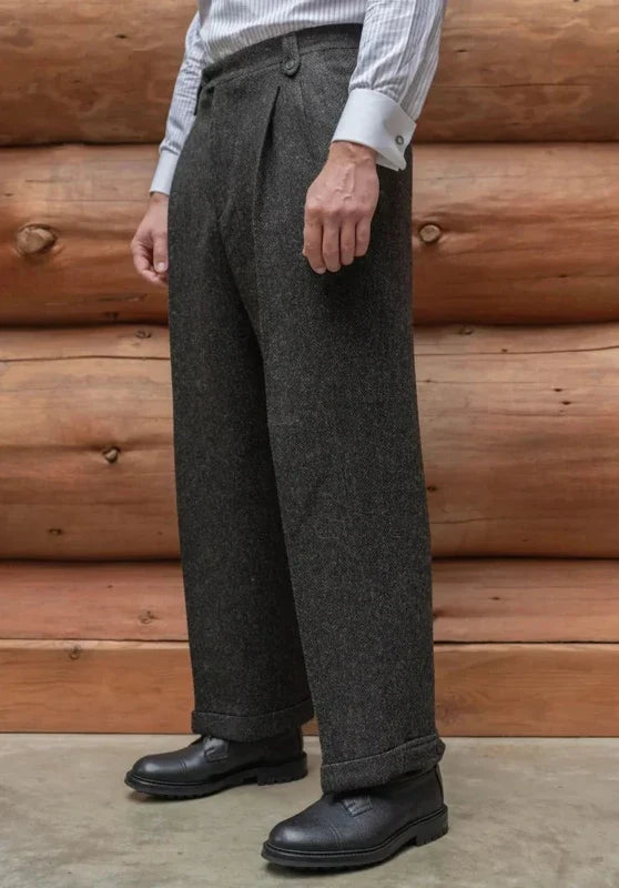 Charcoal gray tweed wool trousers with a straight-leg fit, flat front, and cuffed hems against a rustic log wall.
