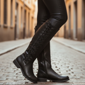 Black knee-high leather boots with lace-up back detailing and low block heels, styled with black leggings in an urban setting.