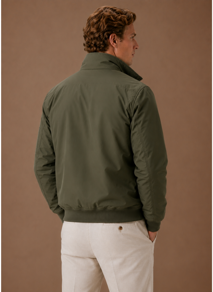 Man wearing an olive green utility bomber jacket over a cream knit sweater with light-colored trousers.