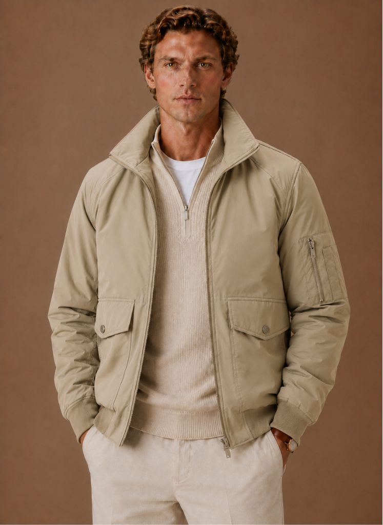 Man wearing a khaki utility bomber jacket with a stand collar, flap pockets, and zip-front closure over a cream knit sweater.