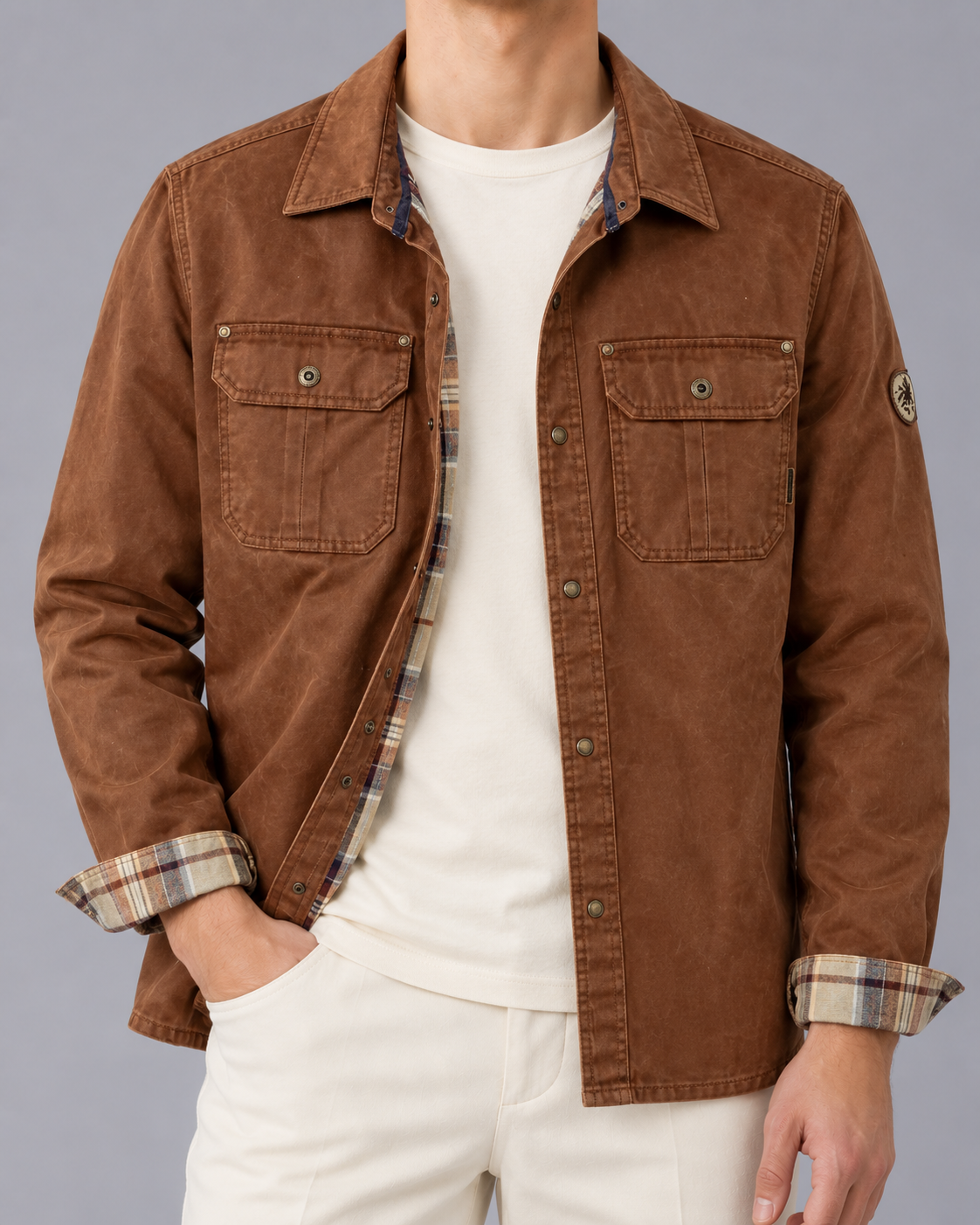Front view of a model wearing the waxed cotton jacket in tobacco brown with snap front buttons, dual flap chest pockets, and plaid cuff trim over a white t-shirt and light trousers.