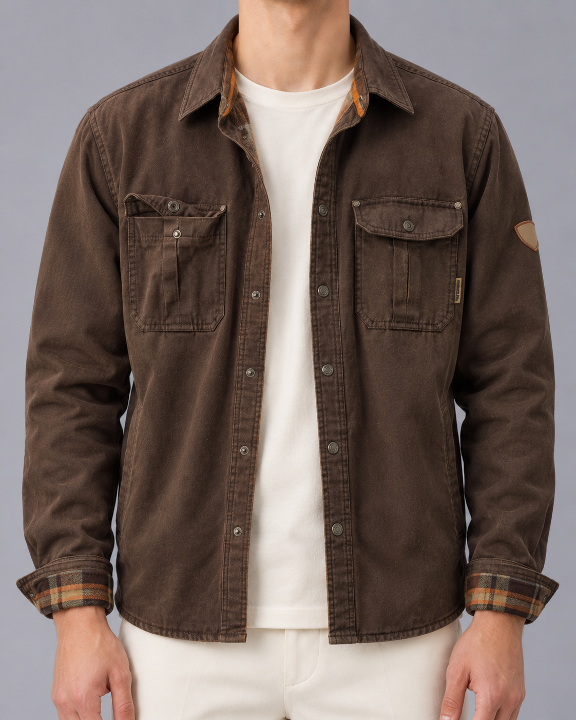 Front view of a model wearing an open dark brown waxed cotton jacket with snap closures, dual flap chest pockets, spread collar, sleeve patch, and subtle plaid-lined cuffs over a white t-shirt.