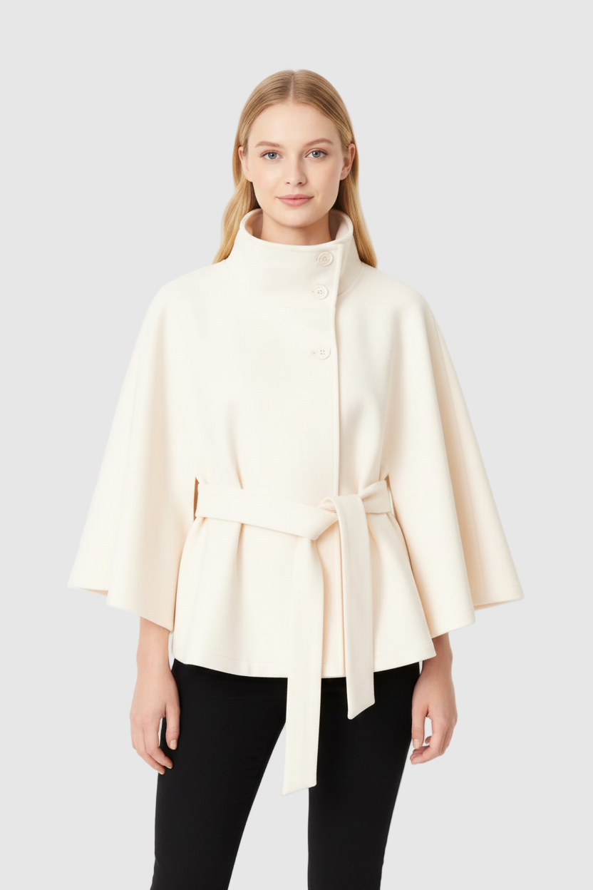 Front view of a model wearing a cream belted wool cape coat with a high stand collar, styled with black trousers against a plain white background.