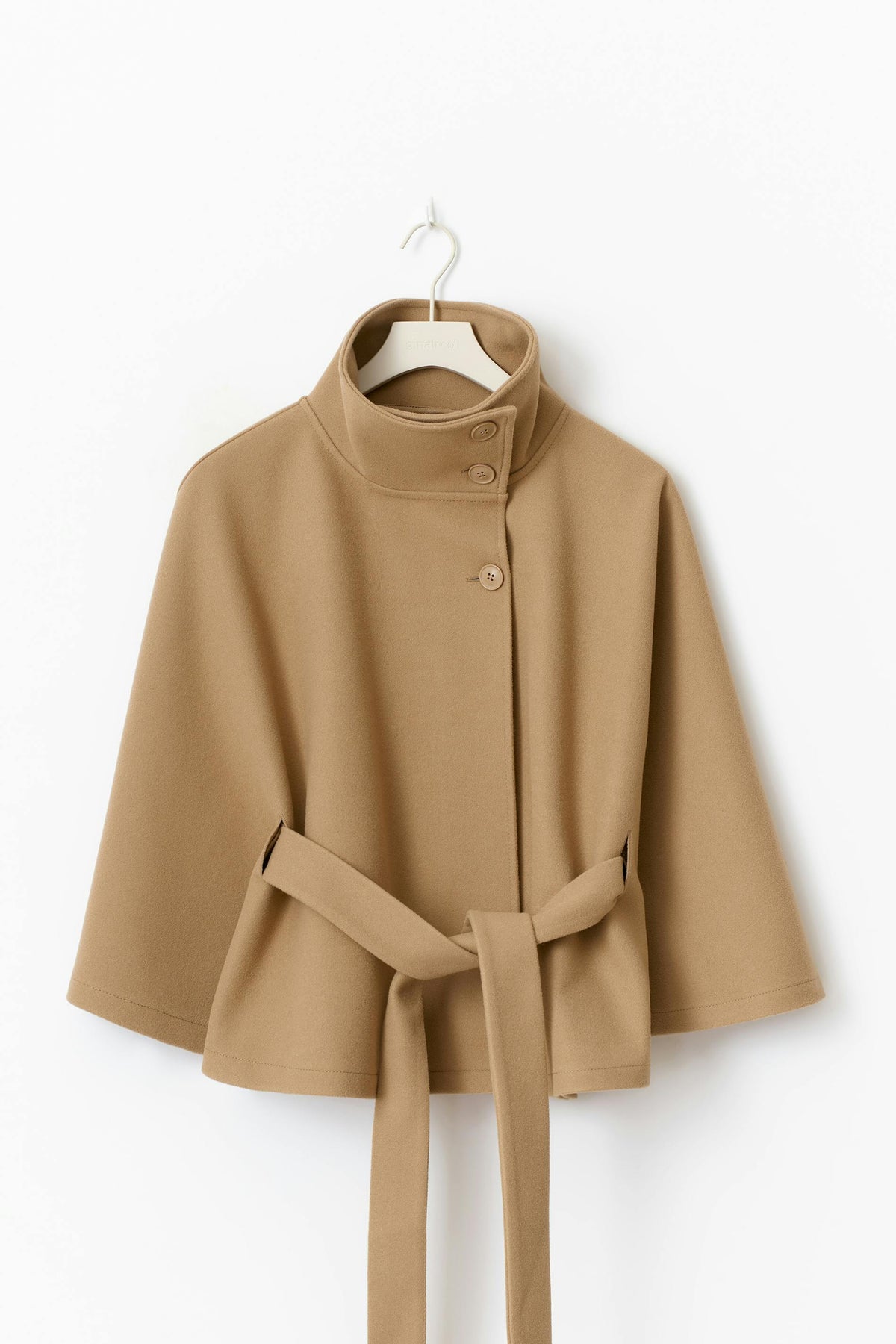 Camel wool cape coat displayed on a hanger featuring a high collar, asymmetric button closure, wide sleeves, and a matching tie waist belt.