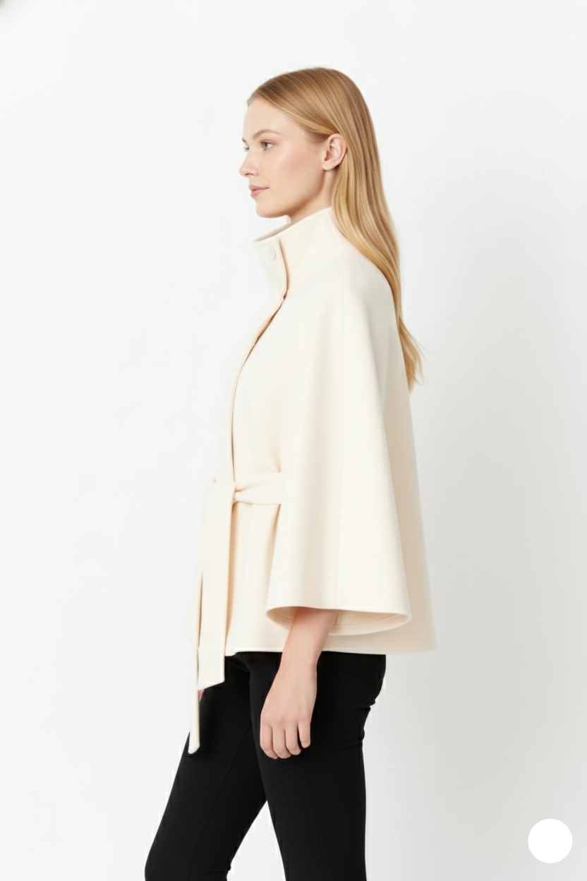 Side profile view of a model wearing a cream belted wool cape coat, showing the flared silhouette and sleeve sleeve cutouts.