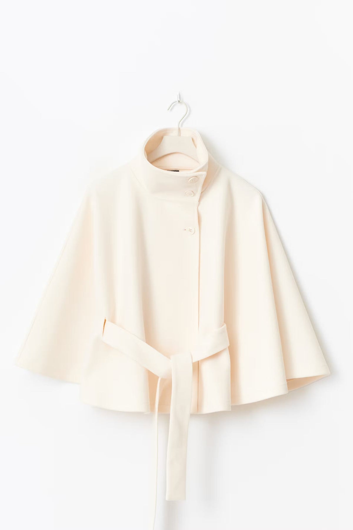 Cream wool-blend cape coat hanging on a hanger featuring a high stand collar, asymmetrical front button closure, wide flared cape sleeves, and a matching tie waist belt.