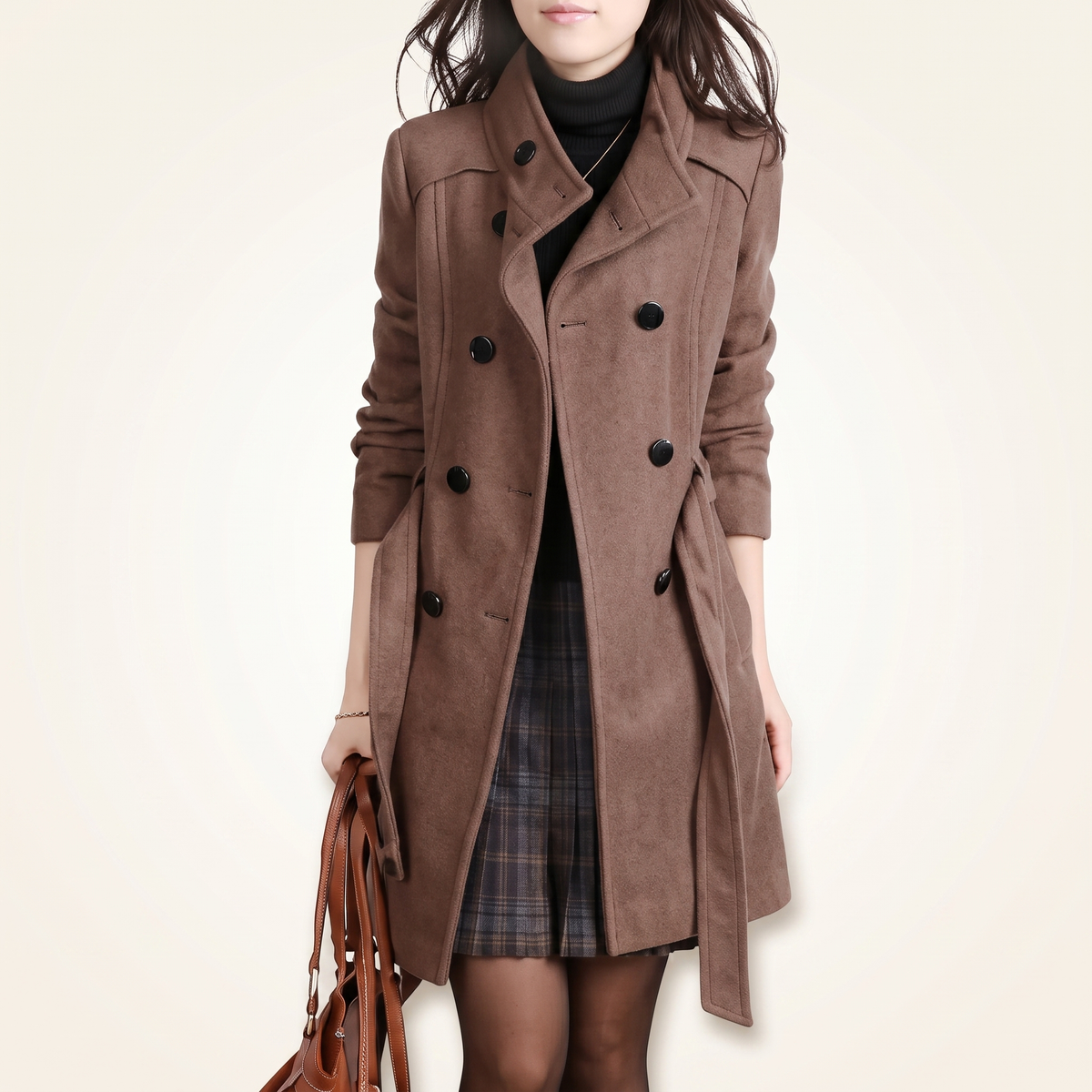 Front view of a woman wearing a brown double-breasted wool coat left open with a tied waist belt, styled over a black turtleneck sweater and plaid skirt