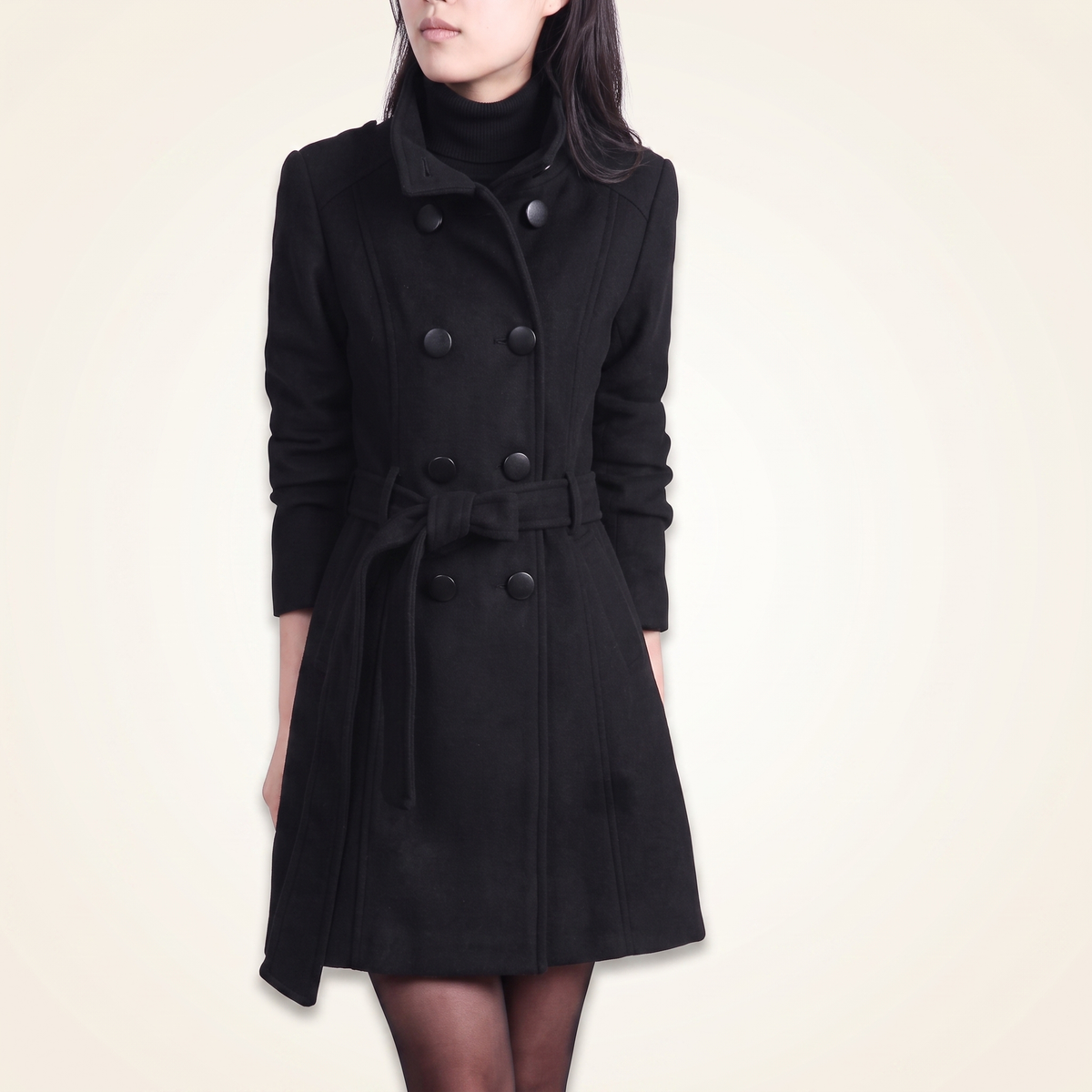 Front view of a woman wearing a black double-breasted wool trench coat buttoned up with a matching tied waist belt and high stand collar