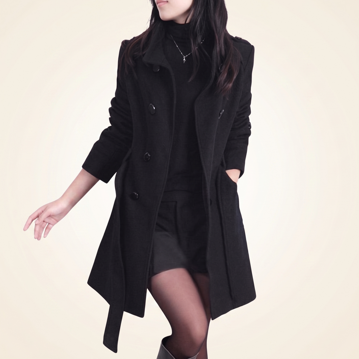 Front view of a woman wearing an unbuttoned black double-breasted wool coat layered over a black top, dark shorts, and tights