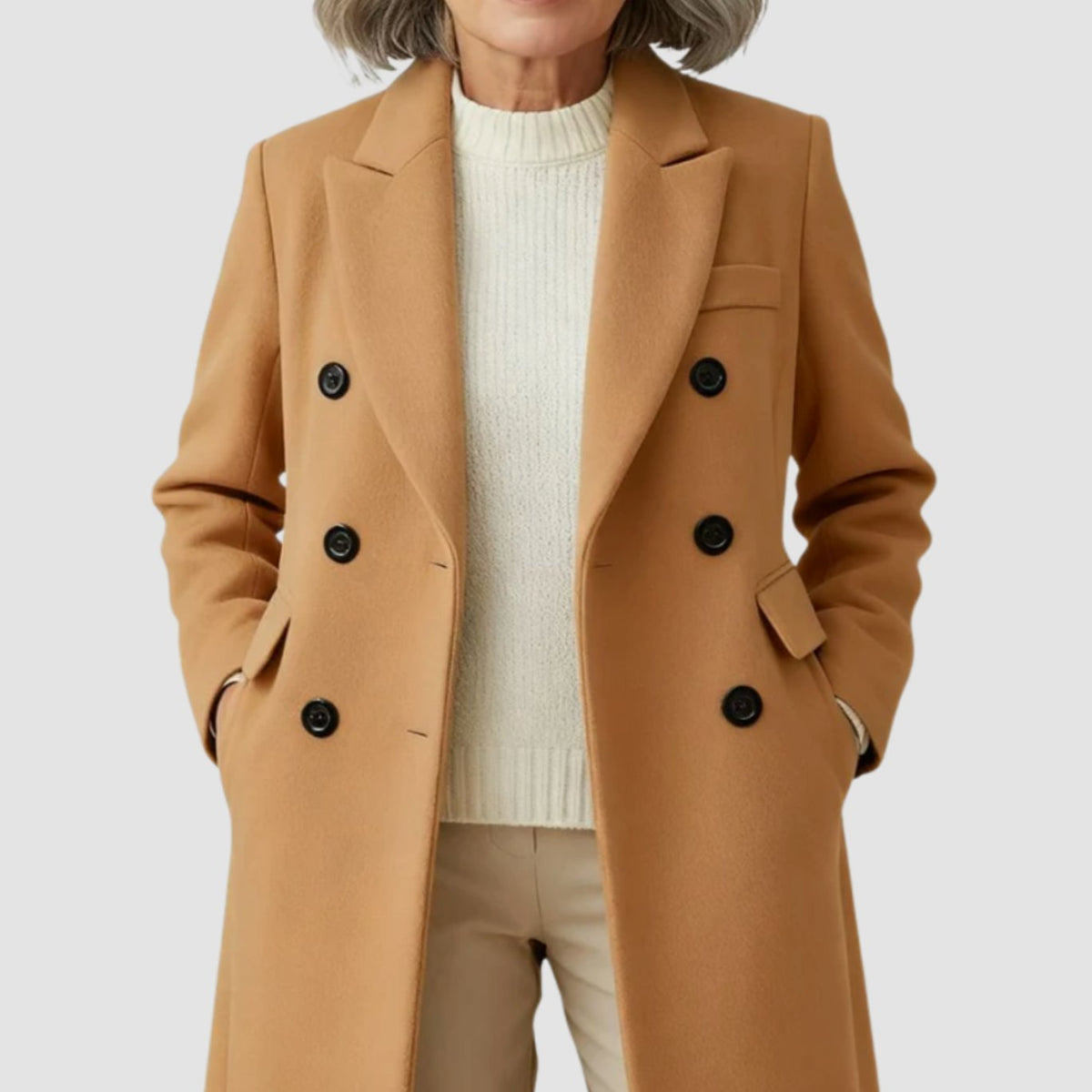 Medium close-up shot of a woman with her hands in the pockets of a camel tan double-breasted coat, styled with a cream ribbed turtleneck sweater and light beige trousers.