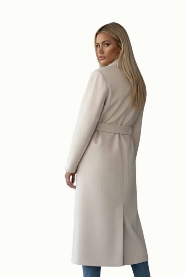 Rear three-quarter view of a woman wearing a light off-white longline double-breasted coat showing the back waist belt structure and central hem vent.