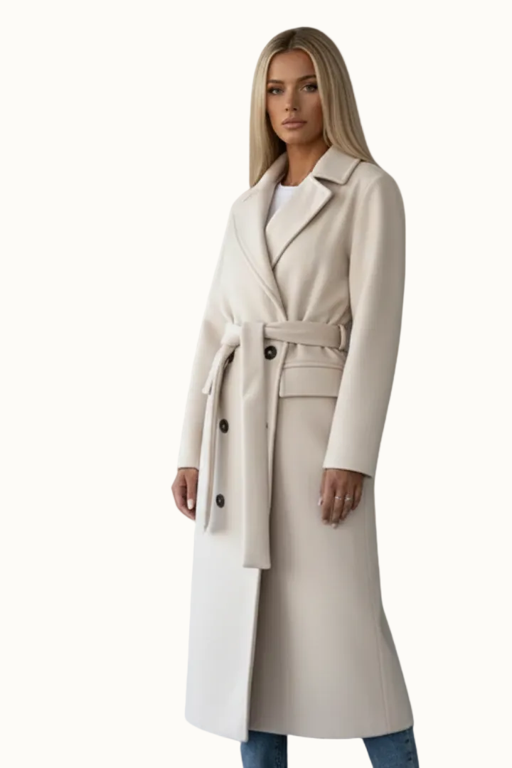 Woman wearing a light off-white longline double-breasted coat featuring notch lapels, dark contrasting buttons, flap pockets, and a tied matching waist belt, styled over blue jeans.