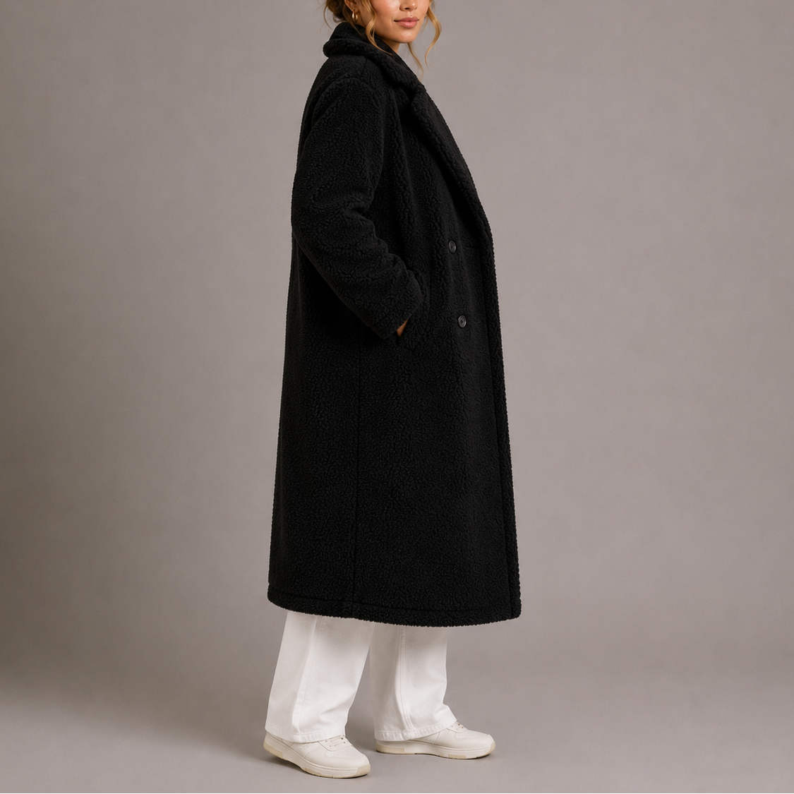 Side profile of a model wearing a black longline teddy coat paired with white trousers and white sneakers.