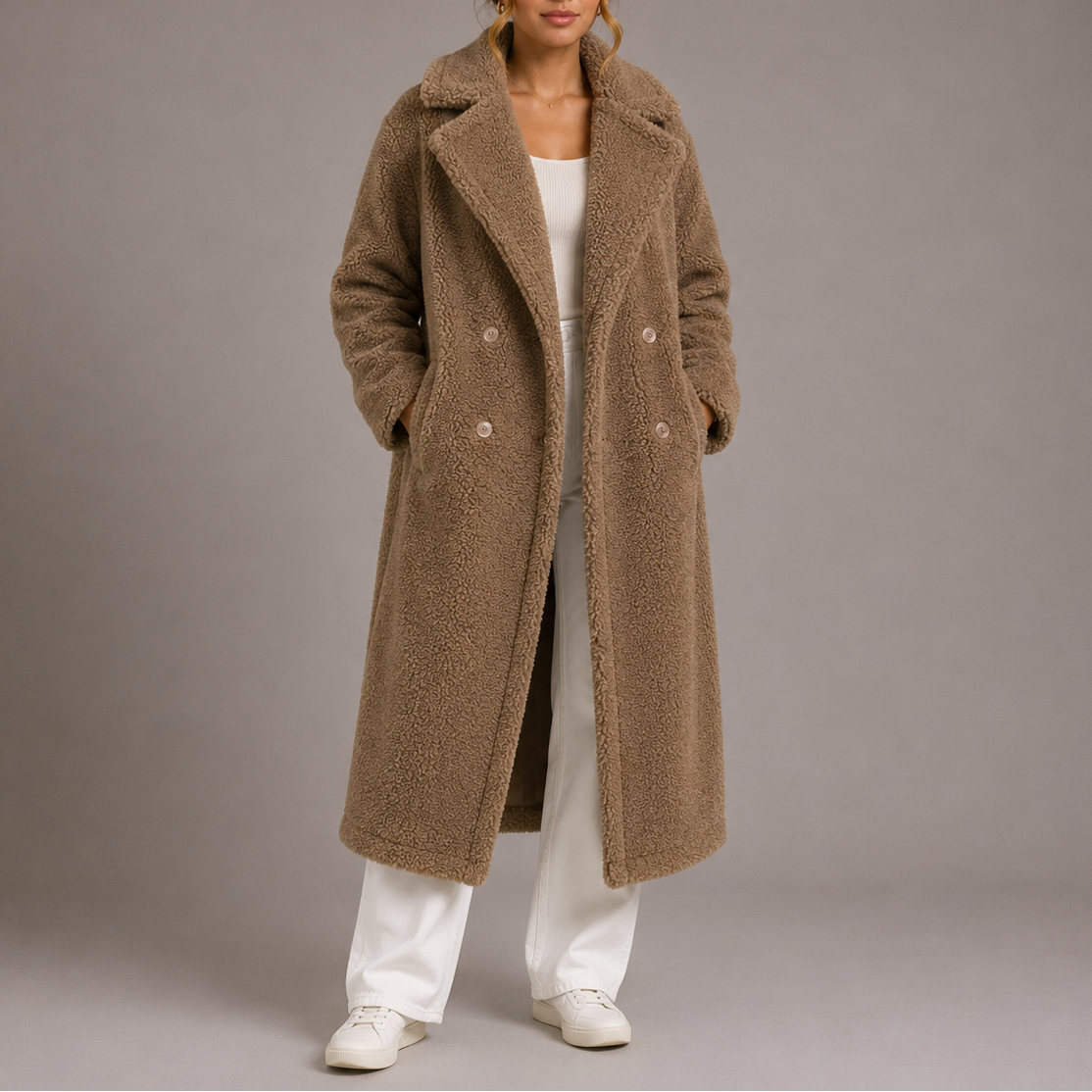 A full-length view of a woman wearing a brown taupe longline teddy coat with notch lapels, layered over a white outfit.