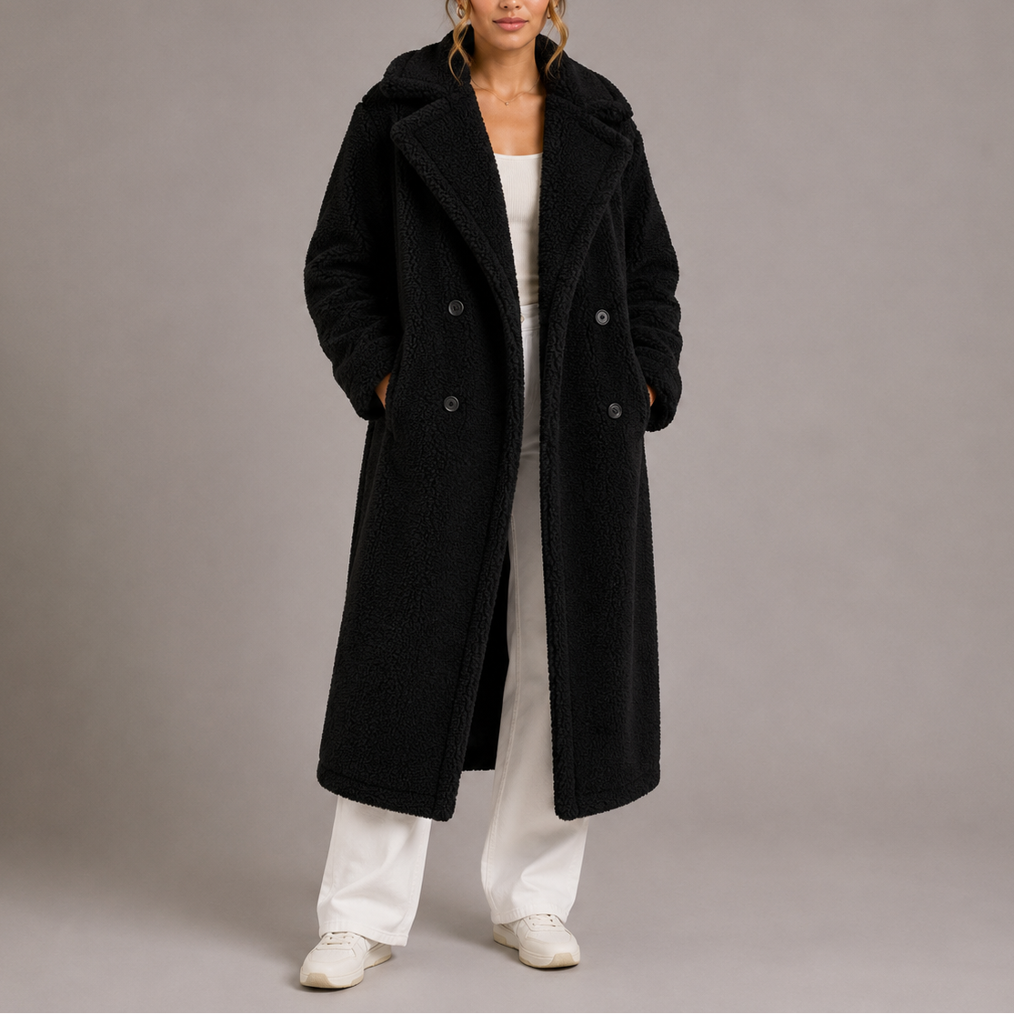 A full-length view of a woman wearing a black longline teddy faux-fur coat with notch lapels and double-breasted buttons, styled over wide-leg white trousers and white sneakers.