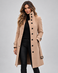 Woman wearing a camel longline wool coat featuring a high funnel neck collar, single-breasted black button closure, buckled cuff straps, and a waist belt with a black buckle, styled over a black turtleneck and dark fitted pants.