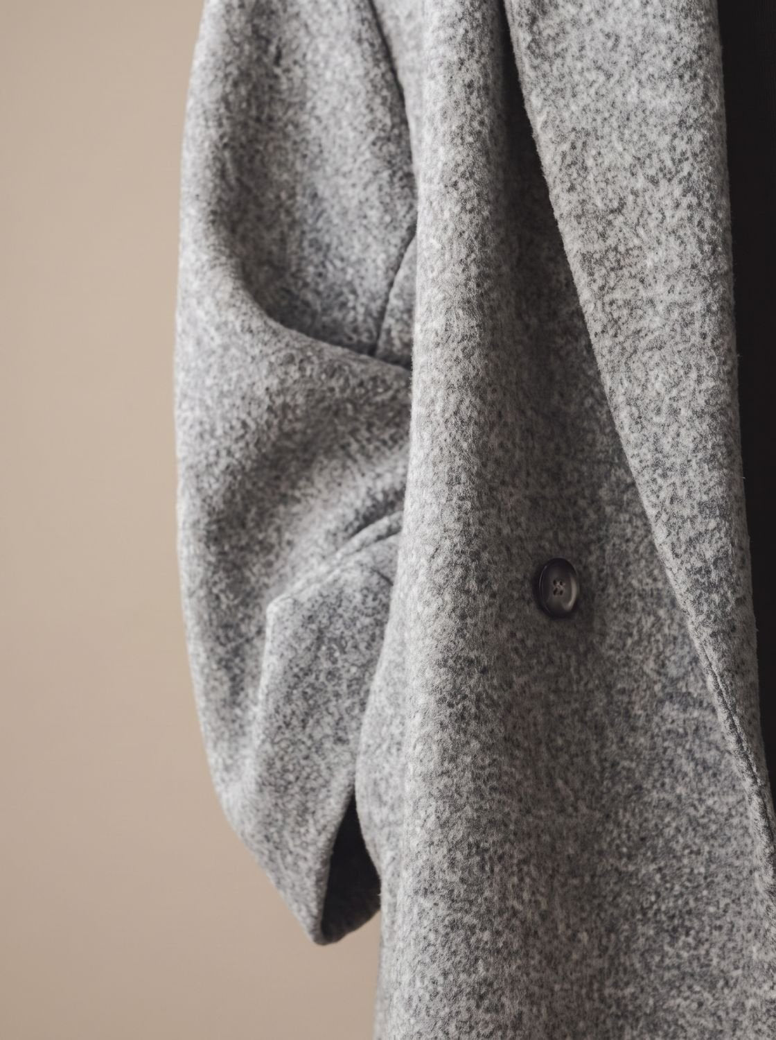 Close-up shot showing the relaxed sleeve, dark button closure, and textured melange wool fabric detail of the light grey overcoat.