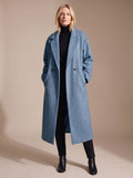 Front full-length shot of a blonde woman wearing a dusty blue oversized double-breasted longline coat styled with a black turtleneck and dark pants.