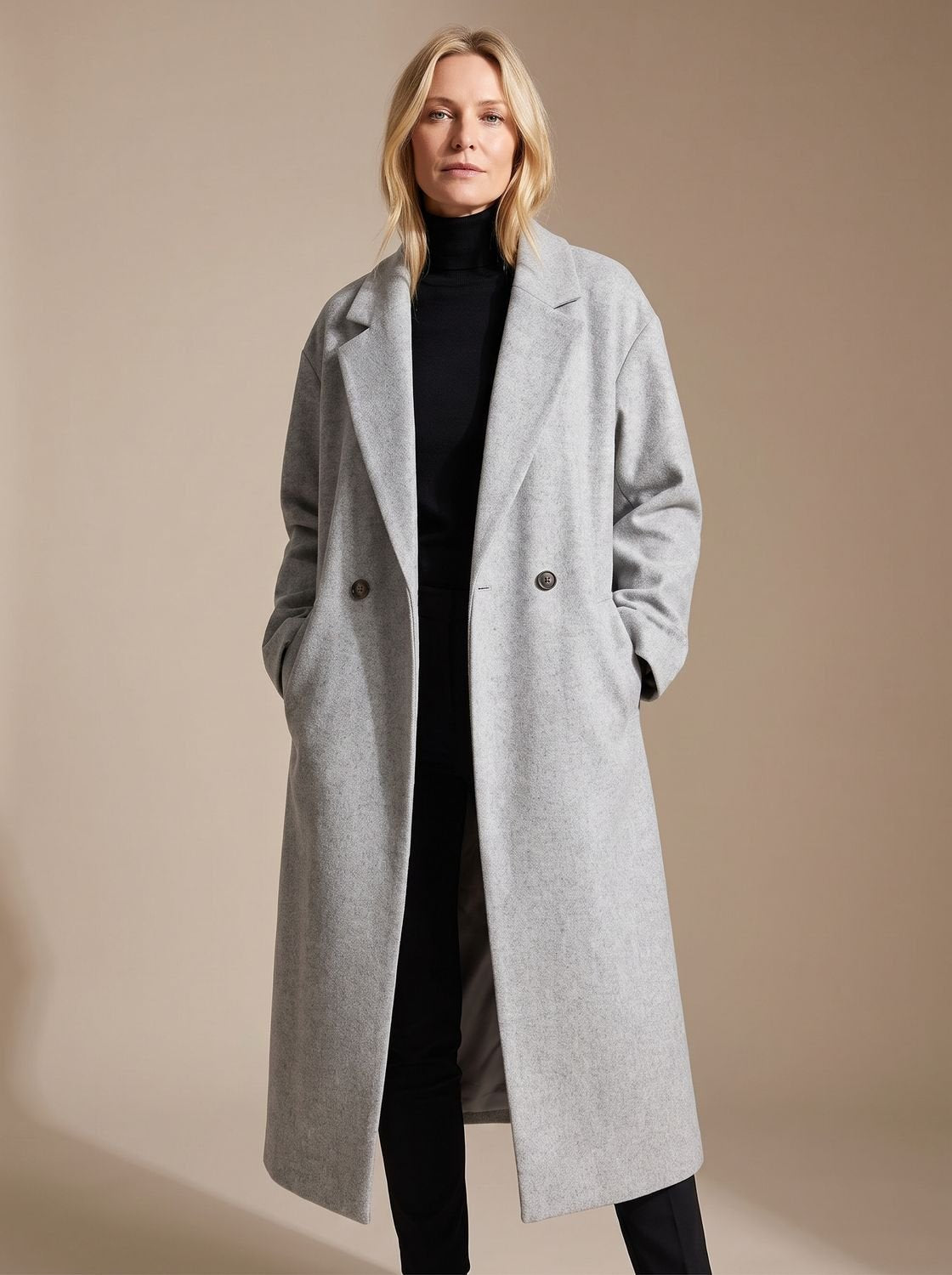Front full-length view of a blonde woman wearing a light grey oversized double-breasted longline coat with wide notch lapels and dropped shoulders, styled over a black turtleneck sweater, dark trousers, and black boots.