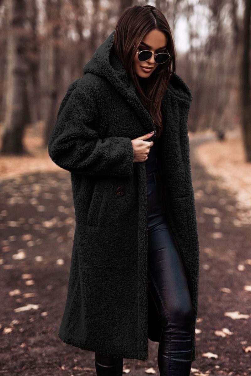 A woman wearing a longline black hooded teddy coat with a button front, styled outdoors over dark trousers and sunglasses.