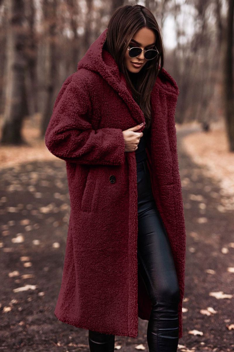 A woman wearing a longline burgundy wine-red hooded teddy coat layered over dark pants and styled with sunglasses.