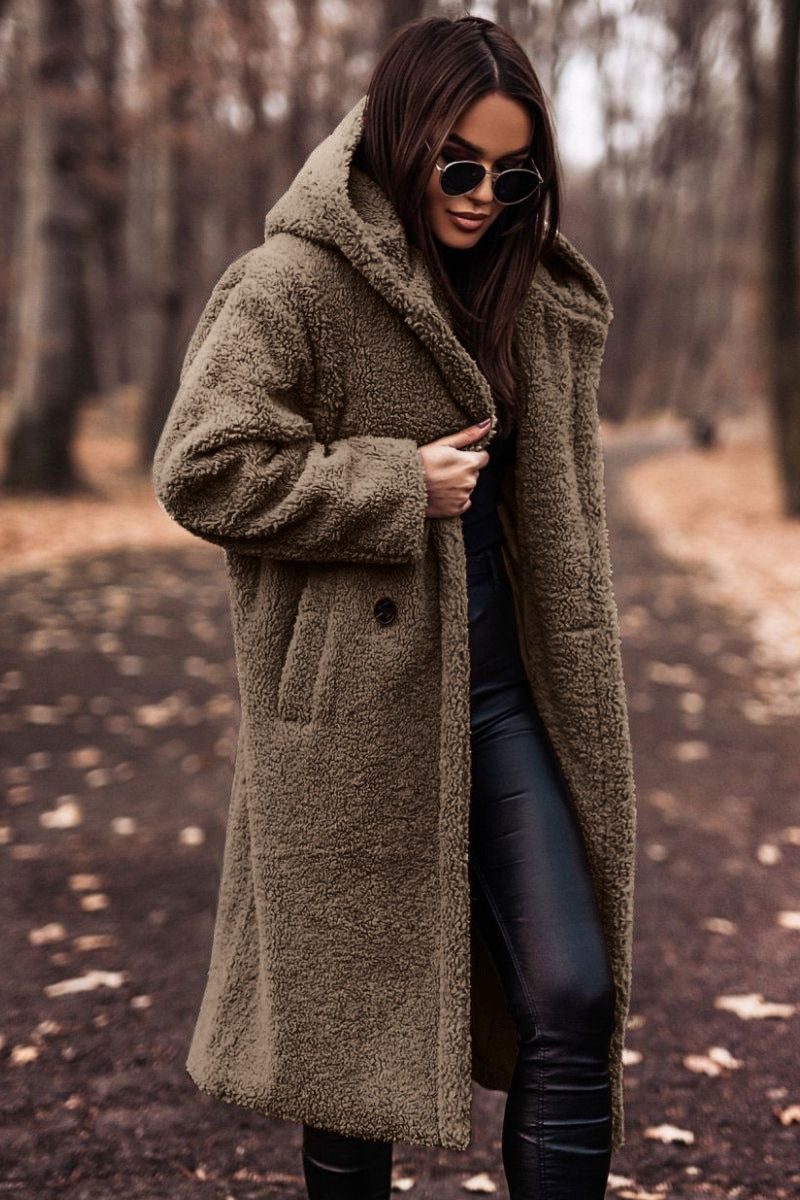 A woman wearing a longline taupe hooded teddy coat worn open over dark pants in an outdoor setting.
