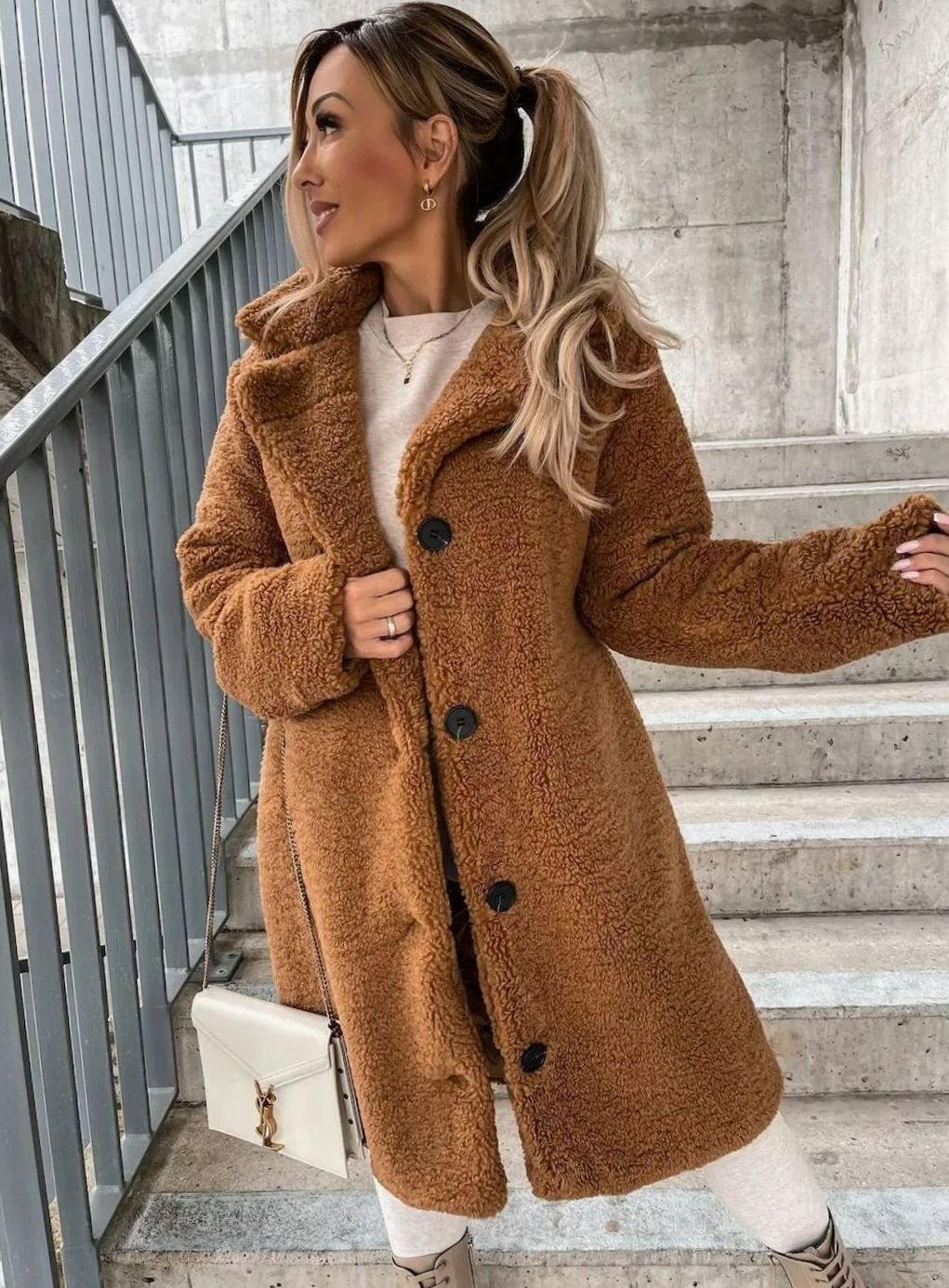 A woman wearing a warm brown midi-length teddy fleece coat with dark front buttons, standing on stairs while holding a white handbag.
