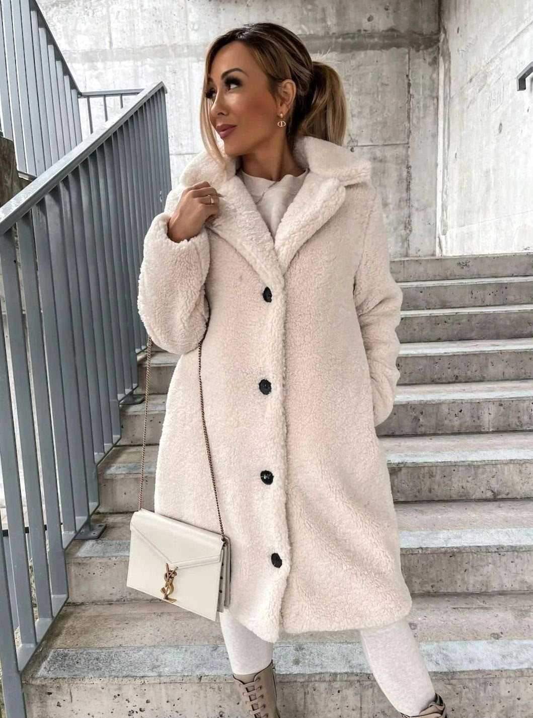 A woman wearing an off-white cream midi teddy coat with contrast buttons, styled with a white crossbody bag, light trousers, and beige boots on a staircase.