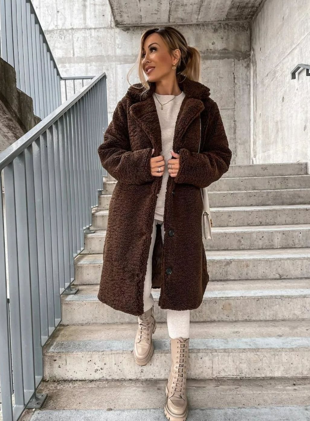 A woman walking down concrete stairs wearing a deep dark brown midi teddy fleece coat paired with a white outfit and light beige boots.