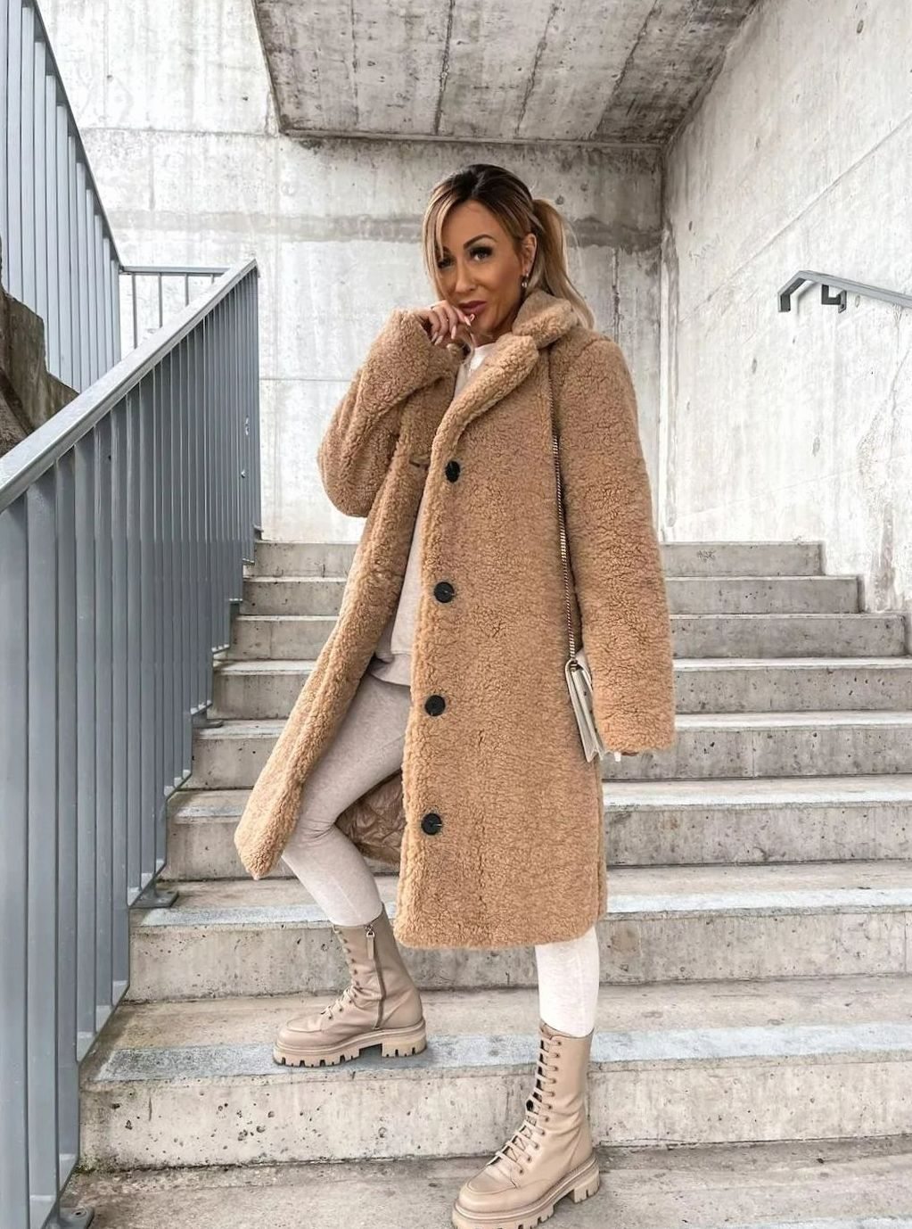 A woman wearing a tan midi-length teddy fleece coat with black button front details, posing on outdoor stairs with neutral trousers and boots.