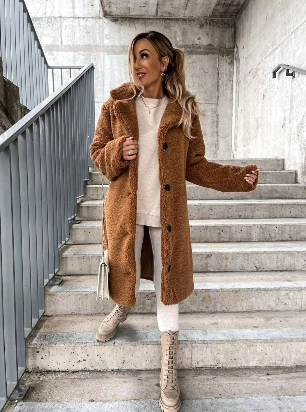 A woman wearing a brown midi teddy fleece coat buttoned down the front, standing on a staircase styled with light pants and boots.