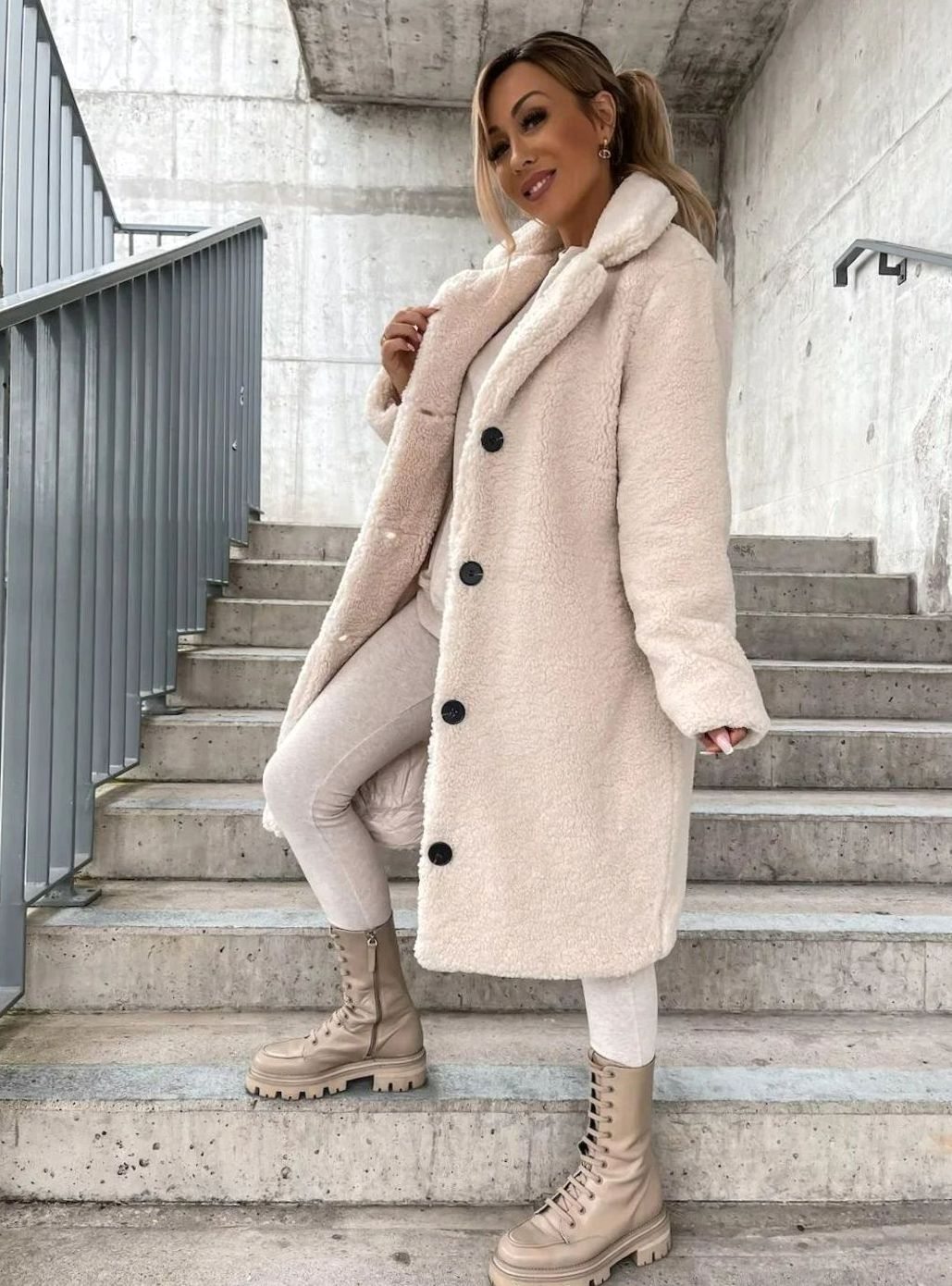 Side view of a woman posing on stairs wearing a cream-colored midi teddy coat with dark buttons and lace-up platform boots.