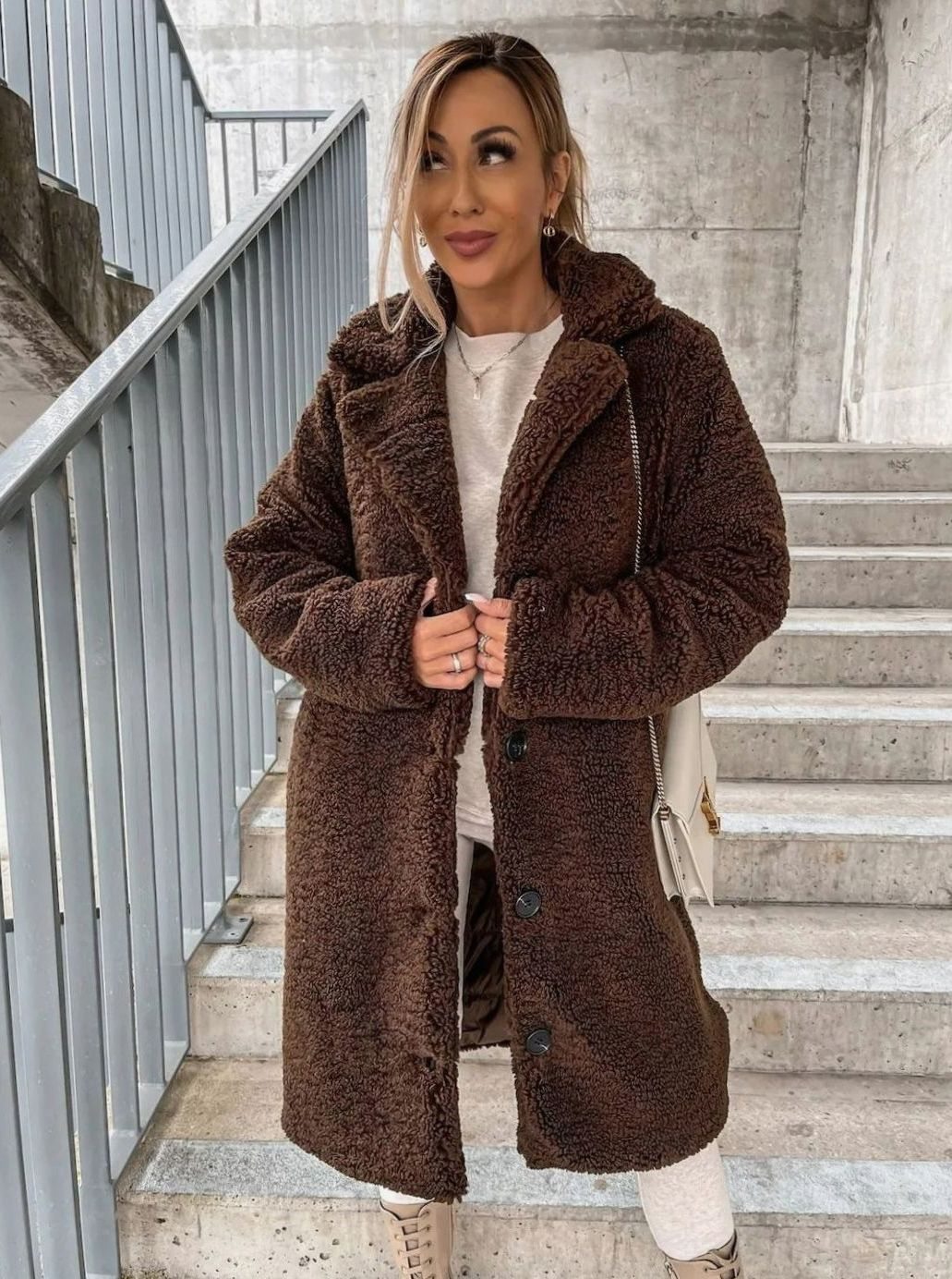 A woman wearing a dark brown oversized midi teddy coat with front buttons, standing on concrete stairs styled with a white top and pants.