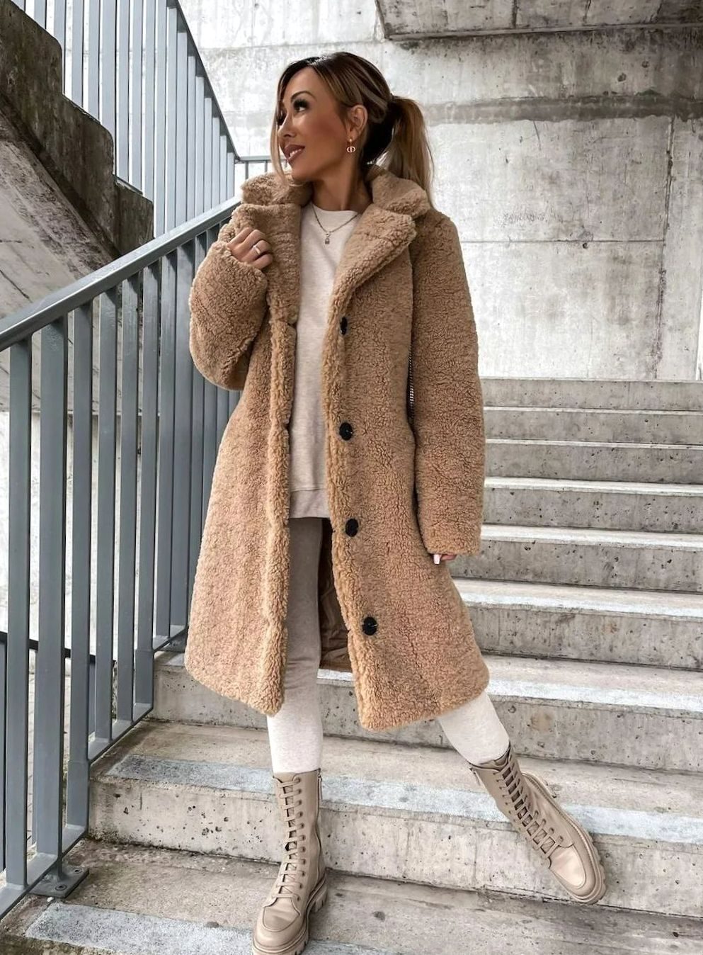 A woman with a ponytail standing on outdoor stairs, wearing a beige midi-length oversized teddy fleece coat with front black buttons, styled over a white top, light pants, and lace-up boots.
