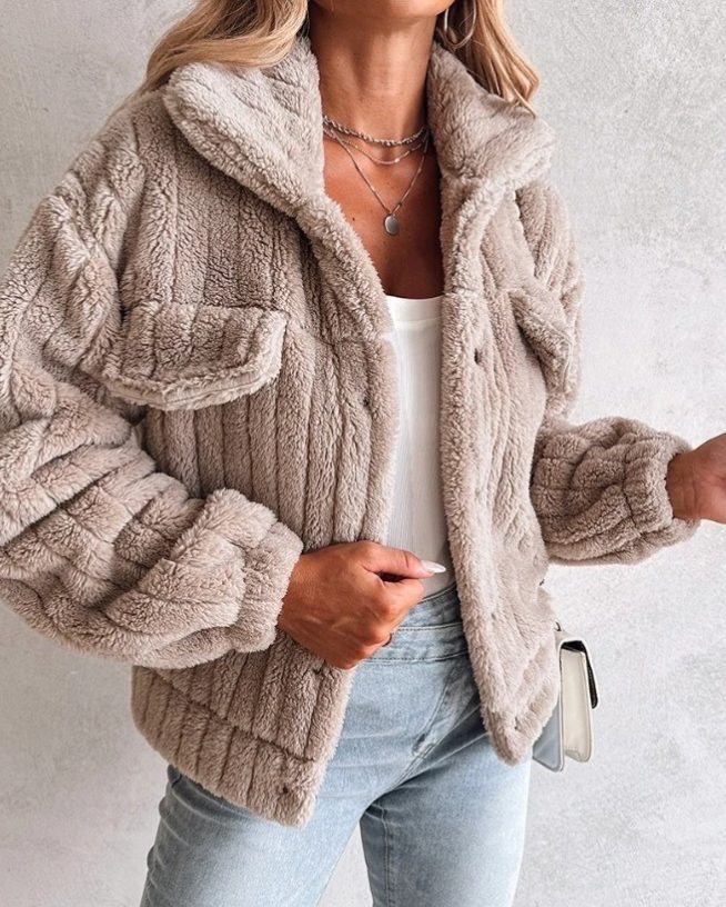 A woman wearing a light beige/taupe oversized teddy fleece jacket with button closure and chest pockets, worn over a white top and light-wash jeans.