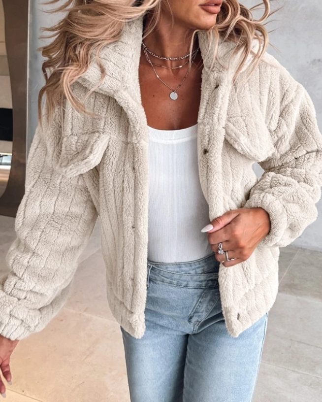 A woman wearing a cream-colored, oversized teddy fleece jacket with a notch collar, flap chest pockets, and button front, styled open over a white ribbed top and light-wash jeans.