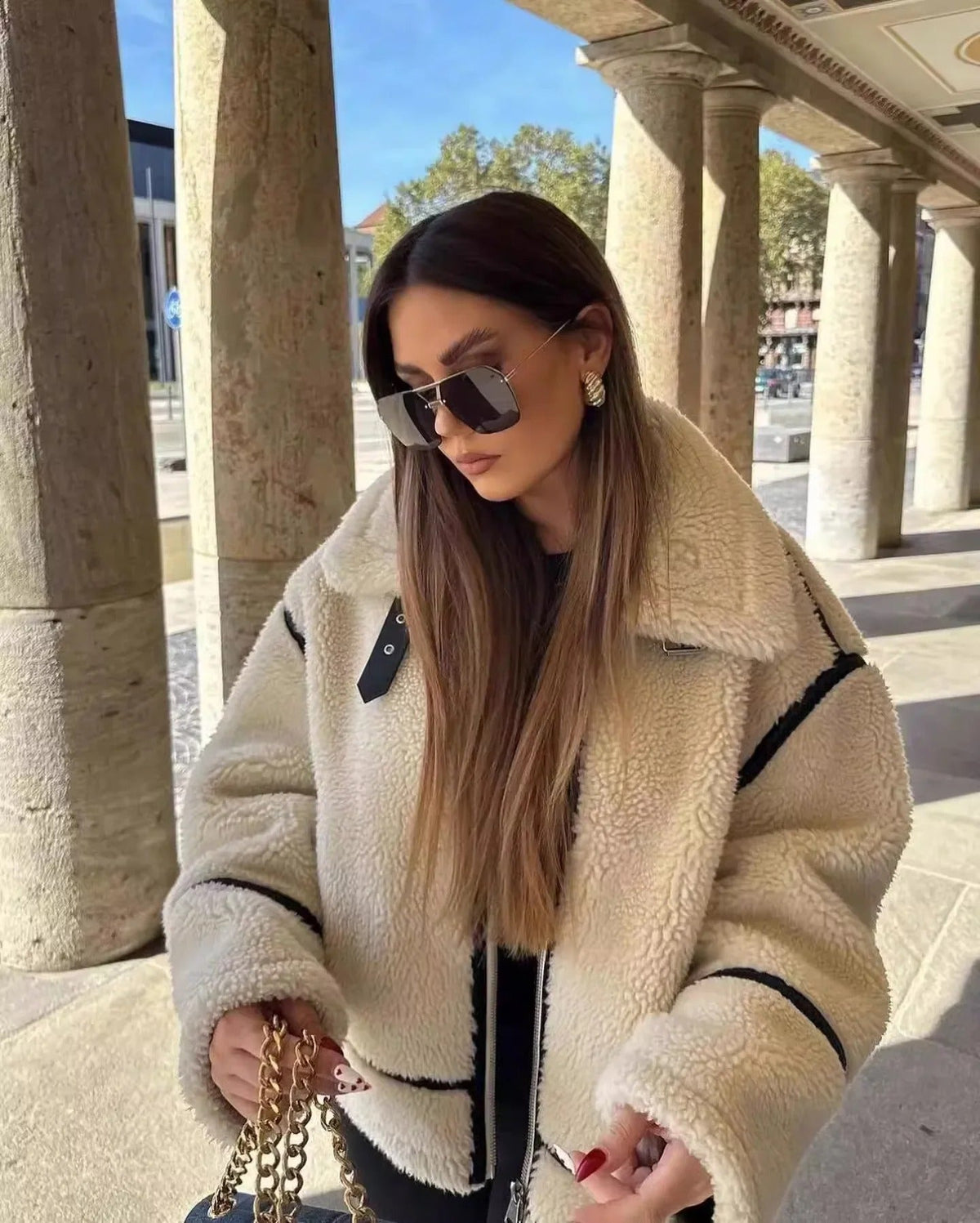 A woman with long hair wearing the oversized cream fleece jacket with black trim outdoors, styled with sunglasses.